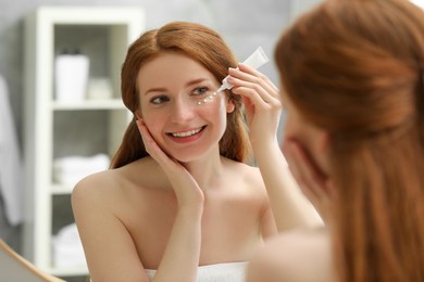 Photo of Smiling woman with freckles applying cream onto her face near mirror in bathroom