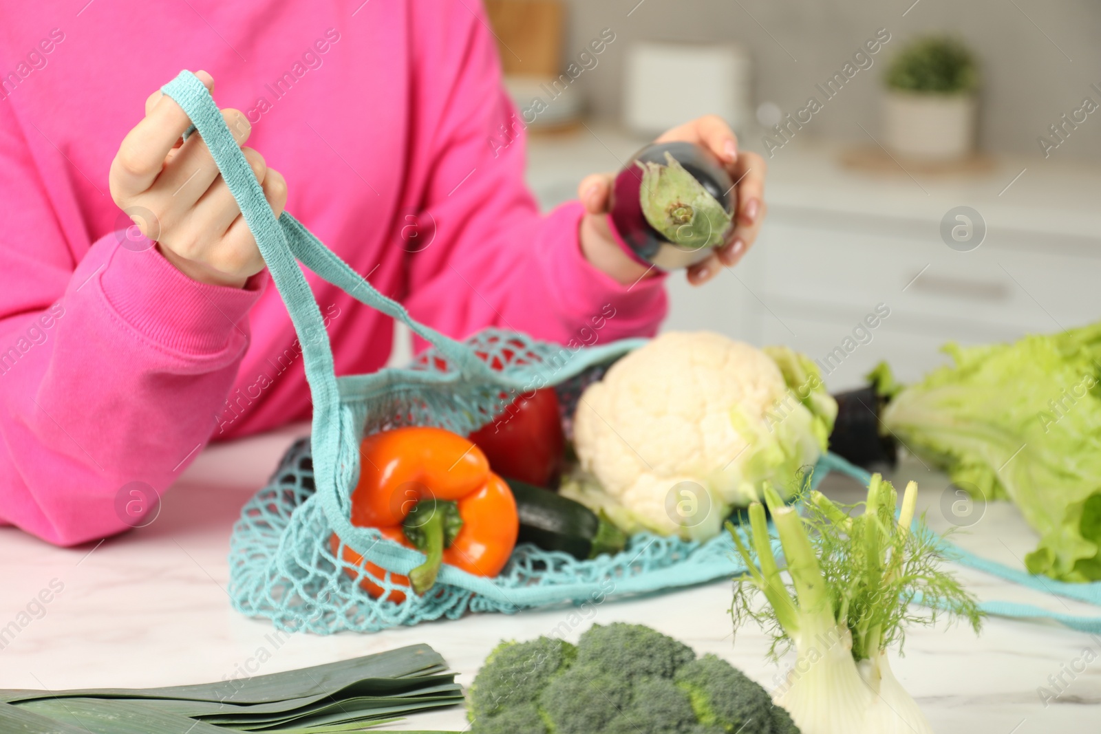 Photo of Woman taking eggplant out from string bag at light marble table, closeup