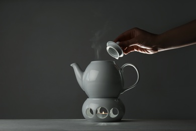 Woman removing lid from ceramic teapot at table against grey background, closeup