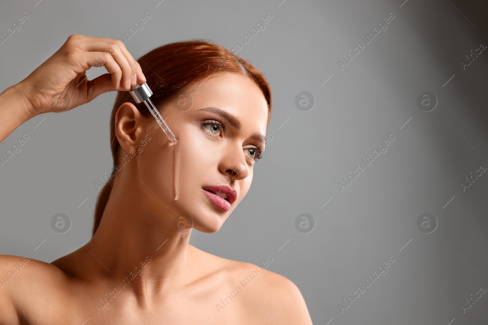 Photo of Beautiful young woman applying cosmetic serum onto her face on grey background, space for text