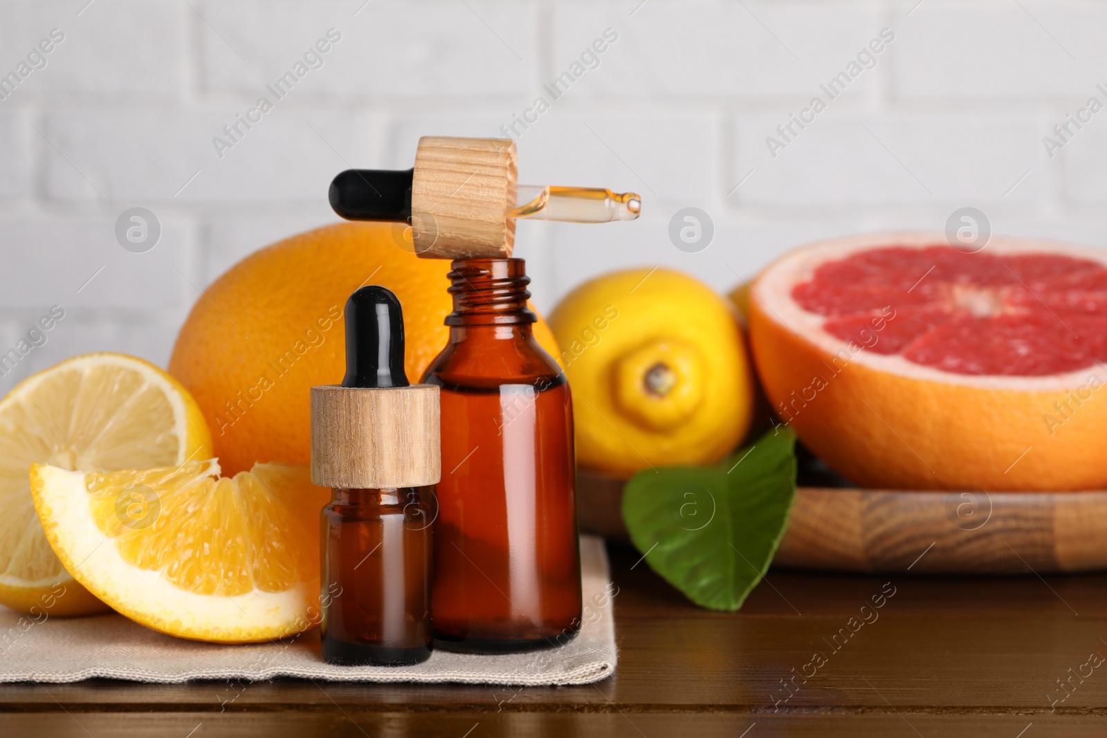 Photo of Bottles of essential oils with different citrus fruits and leaf on wooden table against white brick wall, closeup