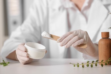 Photo of Dermatologist with jar testing cosmetic product at white table indoors, selective focus