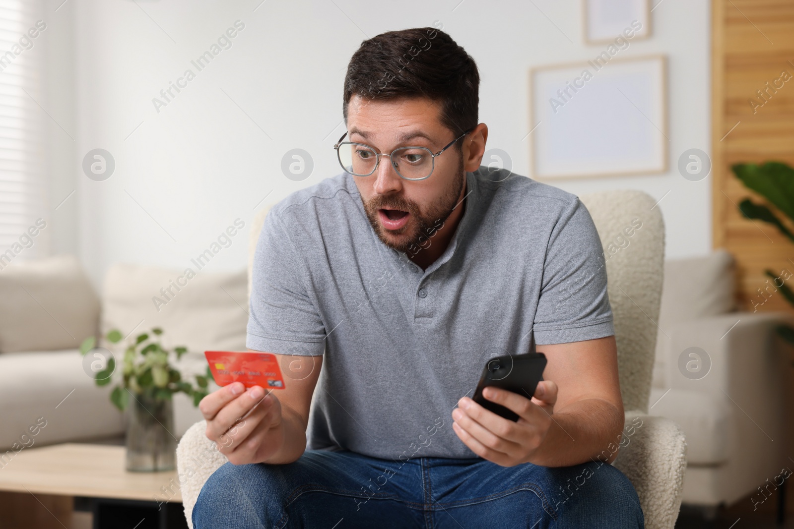 Photo of Scared man with credit card and smartphone in armchair at home. Be careful - fraud