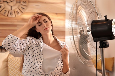 Photo of Woman enjoying air flow from fan on sofa in living room. Summer heat