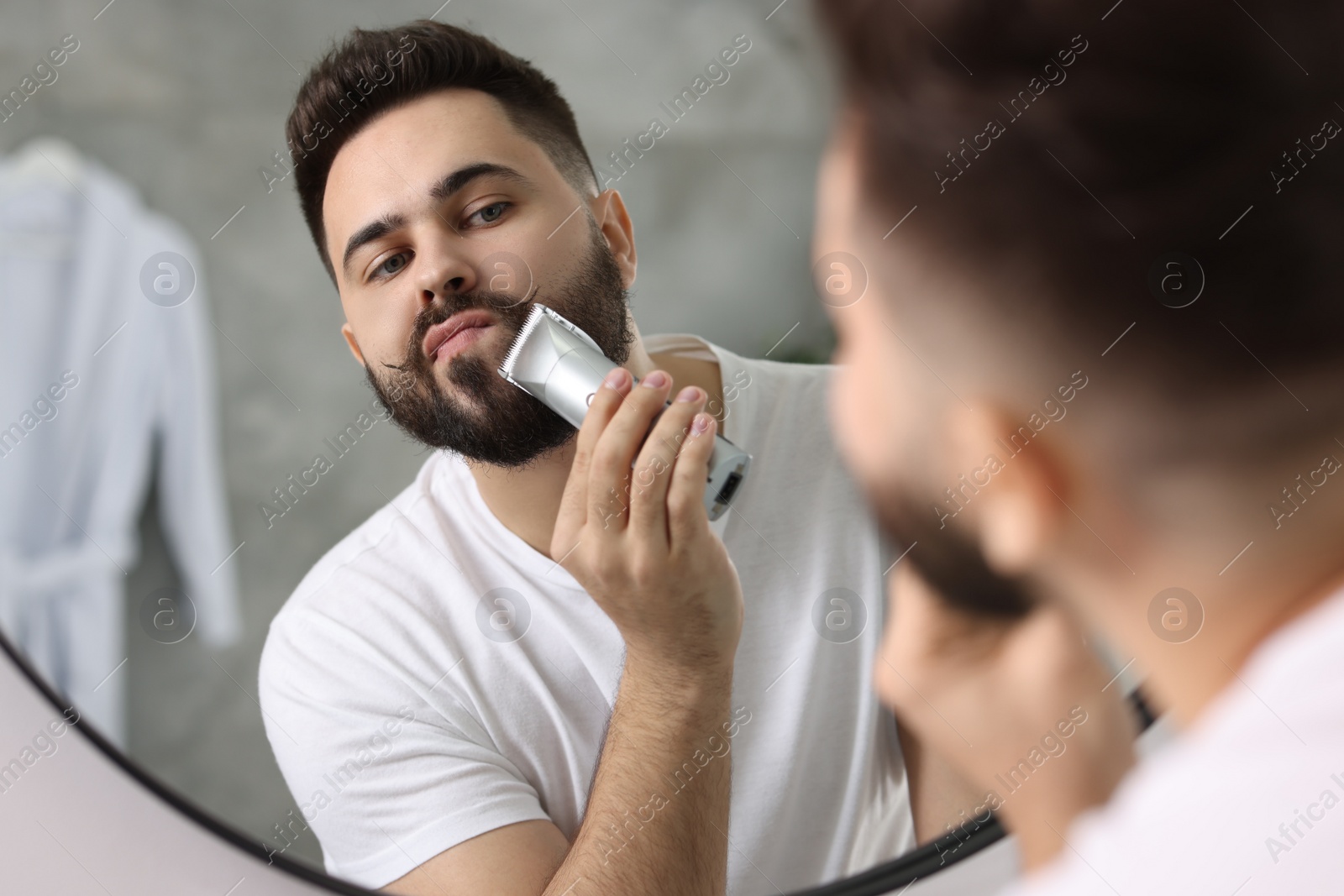 Photo of Handsome young man trimming beard near mirror in bathroom