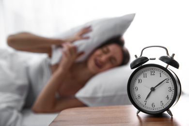 Photo of Young woman covering ears with pillow at home in morning, focus on alarm clock