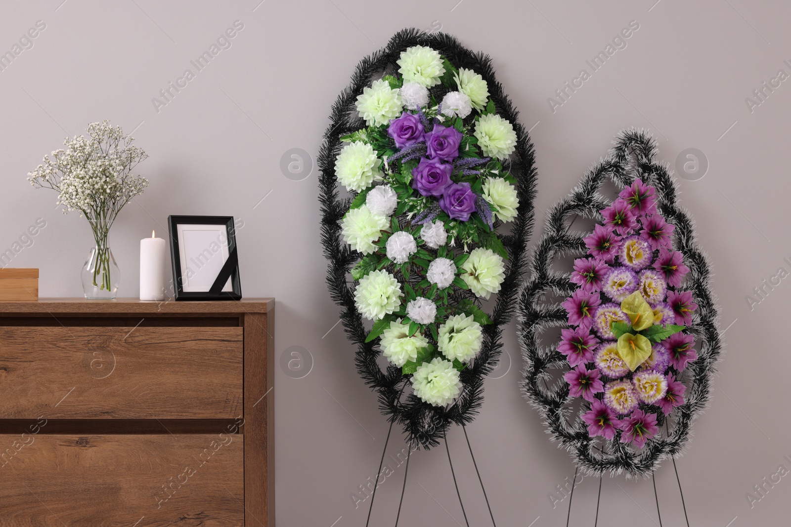 Photo of Wreaths of plastic flowers and frame with black ribbon, burning candle on commode in room. Funeral attributes