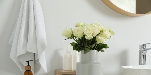 Vase with beautiful white roses and toiletries near sink in bathroom