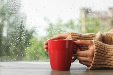 Photo of Woman with cup of hot drink at wooden table near window on rainy day, closeup