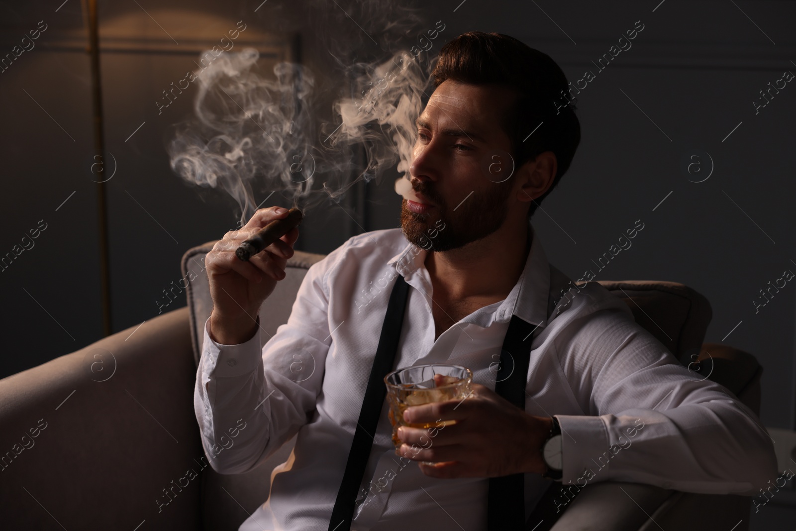 Photo of Handsome man with glass of whiskey smoking cigar in armchair at home