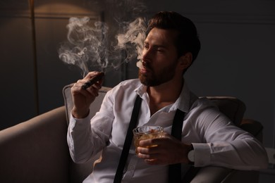 Photo of Handsome man with glass of whiskey smoking cigar in armchair at home