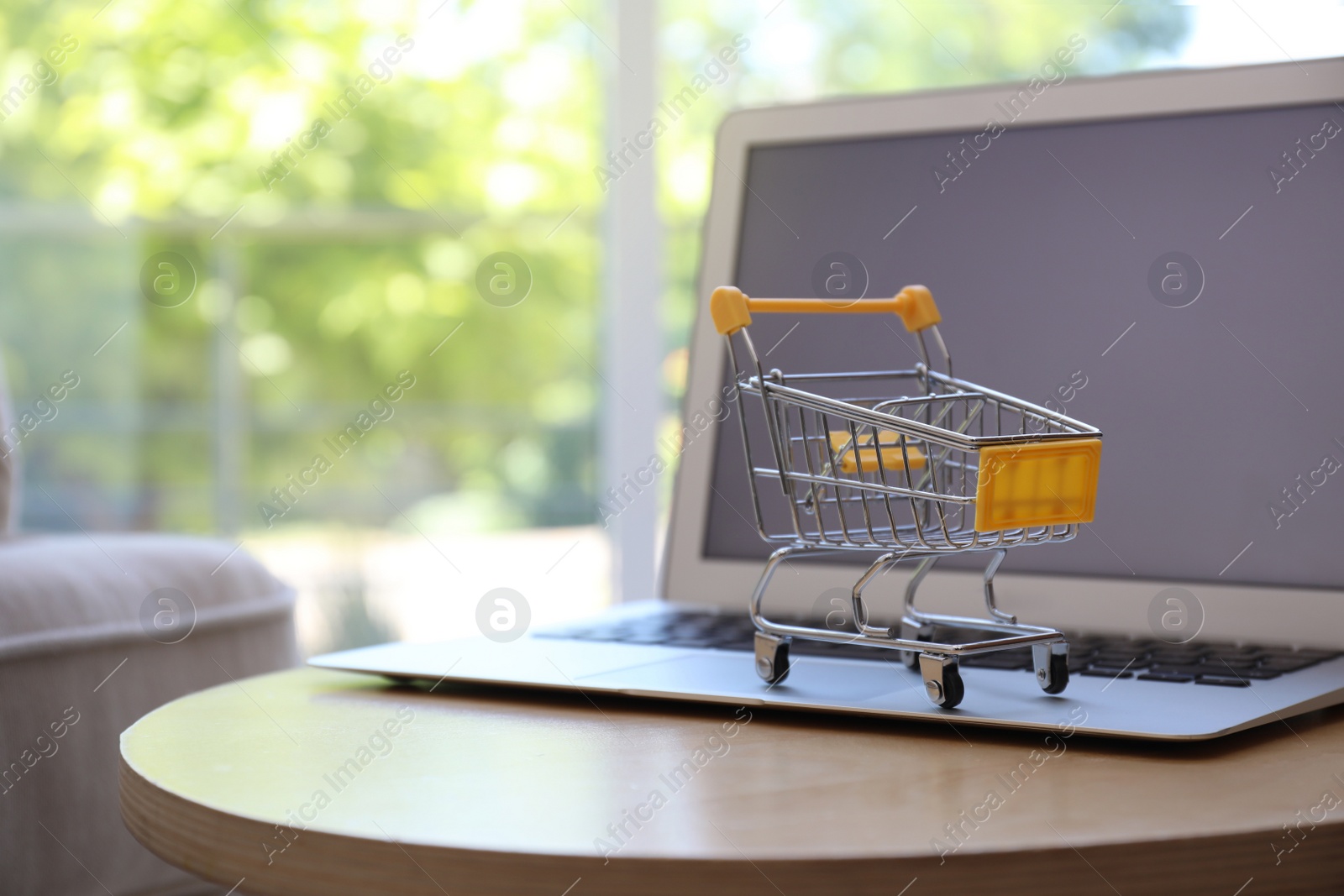 Photo of Internet shopping. Laptop with small cart on table indoors, space for text