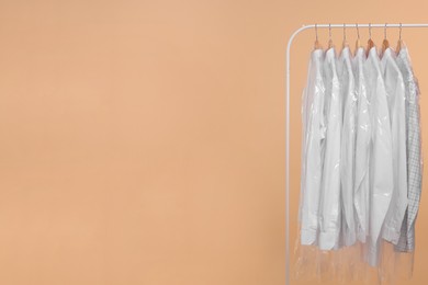 Photo of Dry-cleaning service. Many different clothes in plastic bags hanging on rack against beige background, space for text