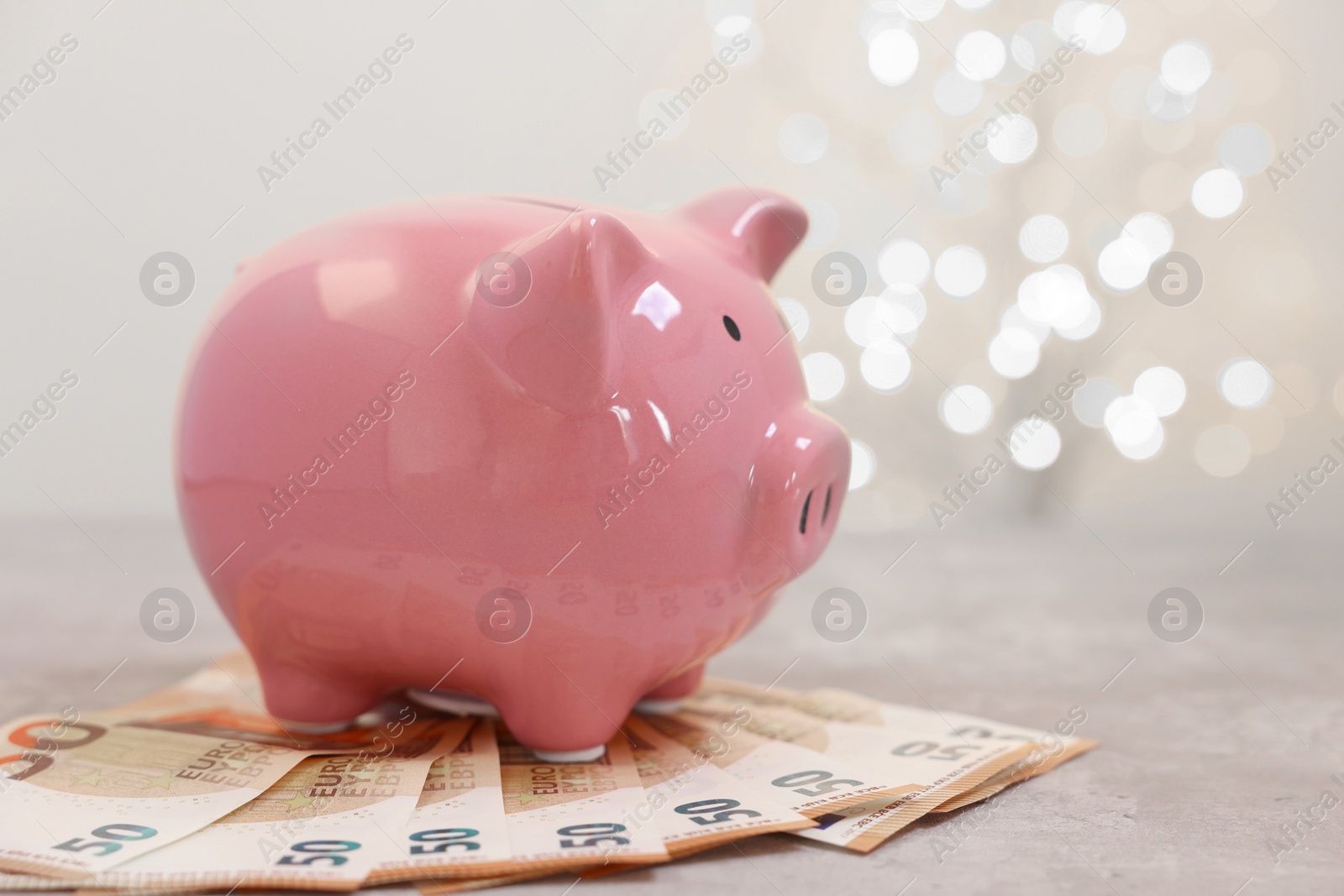 Photo of Piggy bank with euro banknotes on grey table against blurred lights, space for text