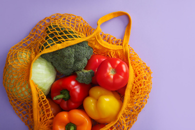 Net bag with vegetables on lilac background, top view