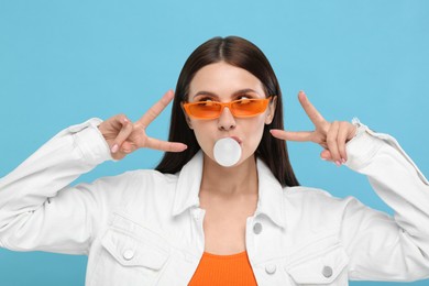 Photo of Beautiful woman in sunglasses blowing bubble gum and gesturing on light blue background