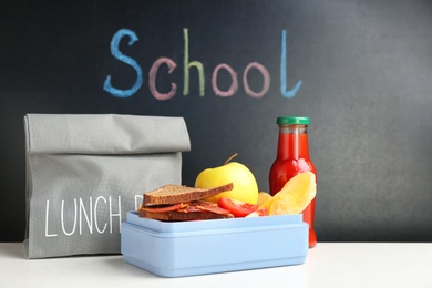 Photo of Appetizing food in lunch box and bag on table near chalkboard with word SCHOOL