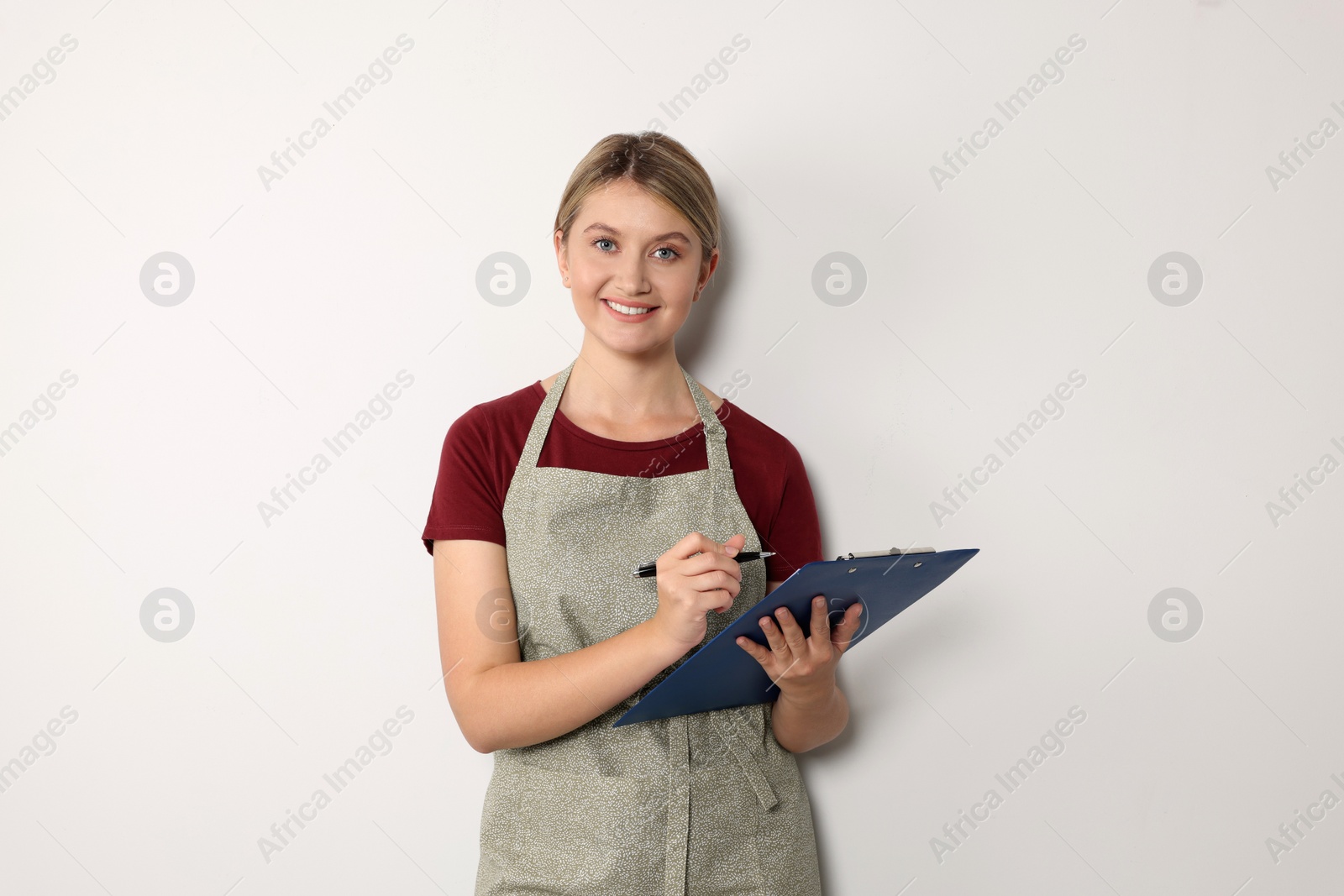 Photo of Beautiful young woman in clean apron with clipboard on light grey background