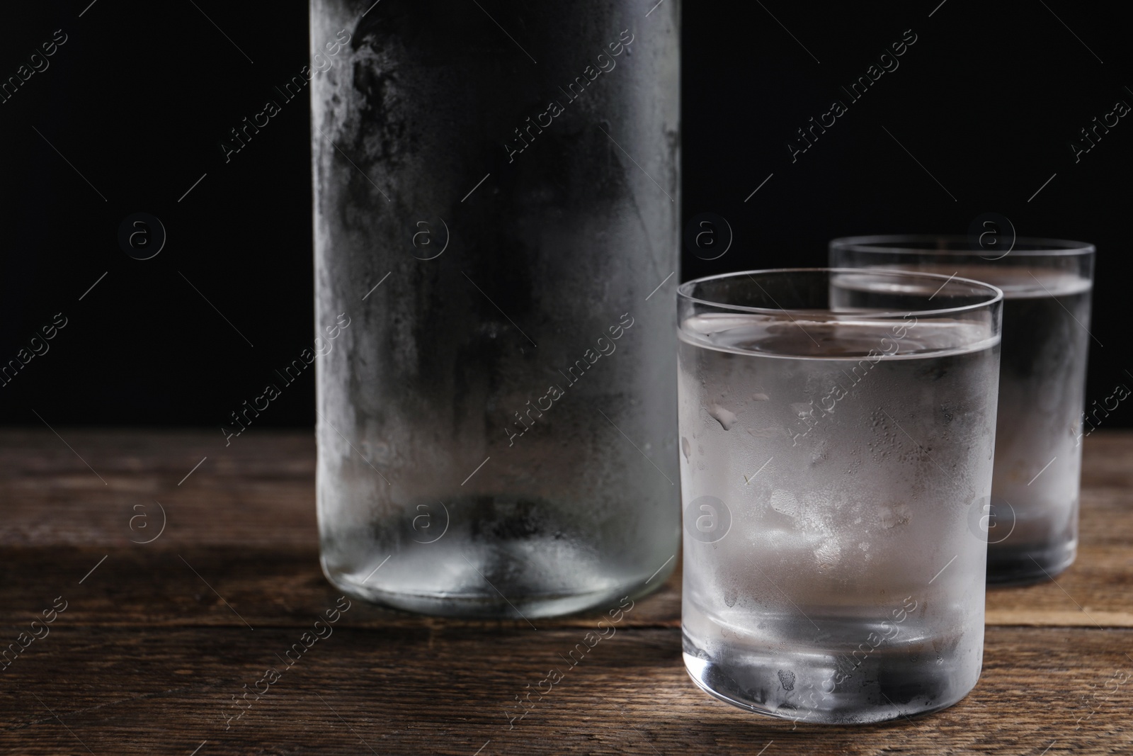 Photo of Bottle of vodka and shot glasses on wooden table against black background, closeup