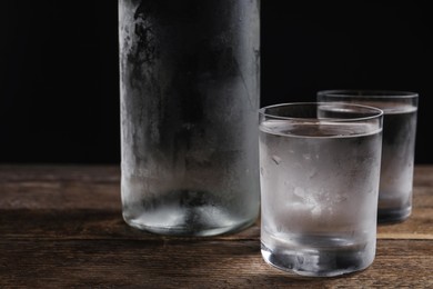 Photo of Bottle of vodka and shot glasses on wooden table against black background, closeup