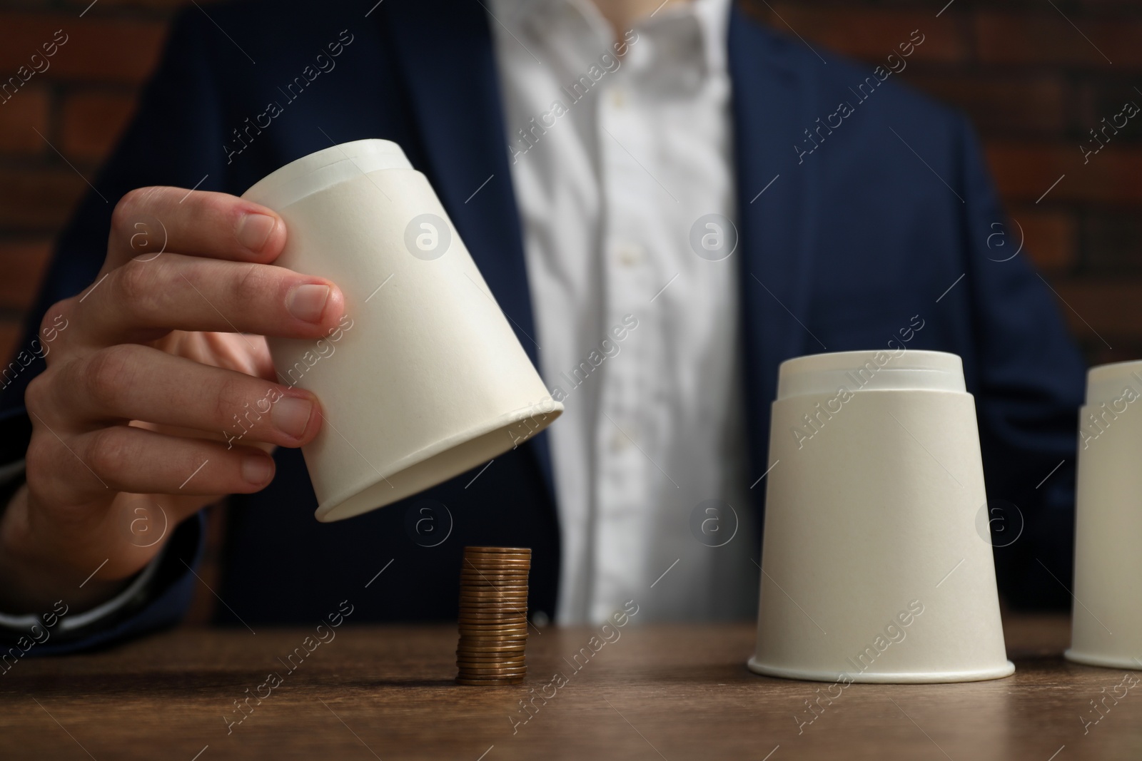 Photo of Shell game. Man showing stack of coins under cup at wooden table, closeup