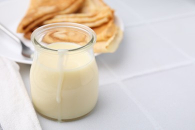 Photo of Tasty condensed milk in jar and crepes on white tiled table, closeup. Space for text