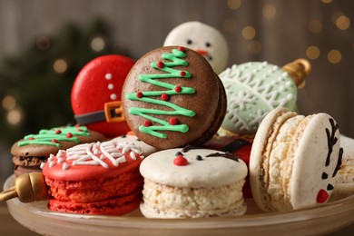 Photo of Beautifully decorated Christmas macarons on dish against blurred festive lights, closeup