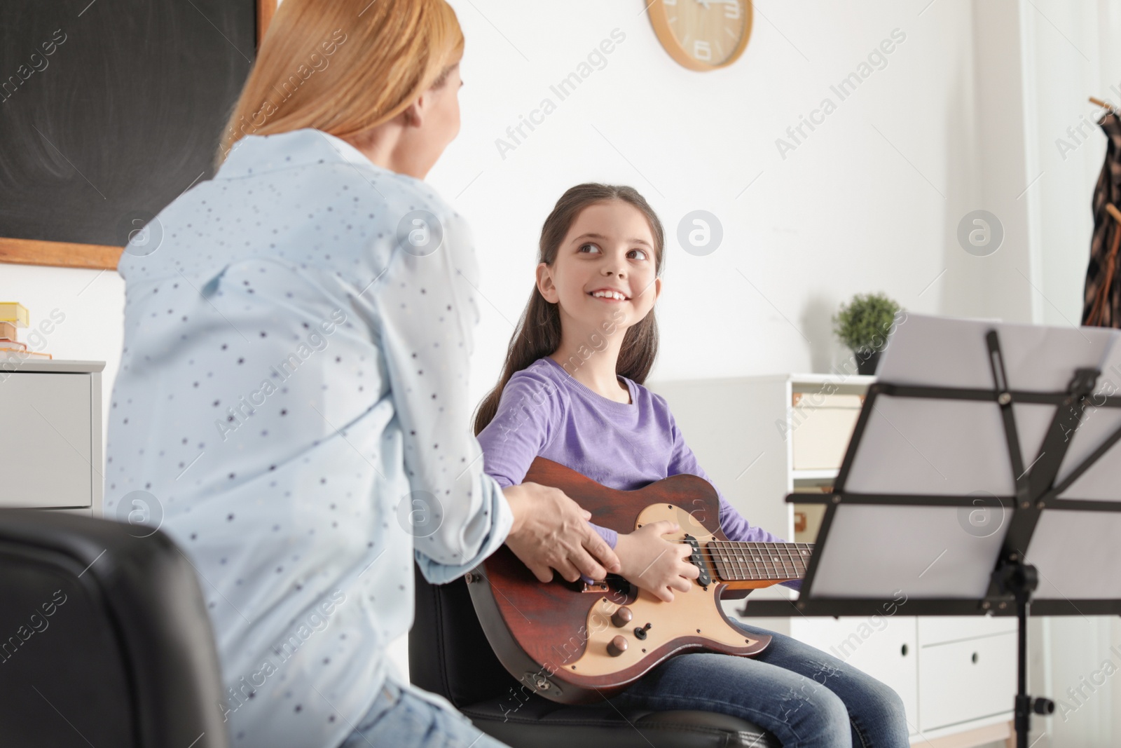 Photo of Little girl playing guitar with her teacher at music lesson. Learning notes