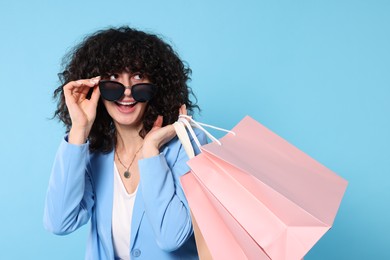 Happy young woman with shopping bags and stylish sunglasses on light blue background