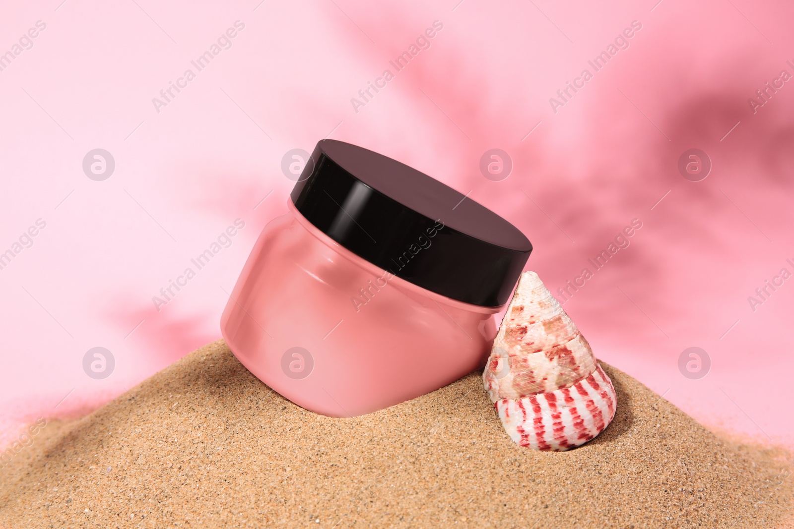 Photo of Jar of body cream and seashell on sand against pink background