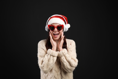 Young woman in Santa hat listening to Christmas music on black background