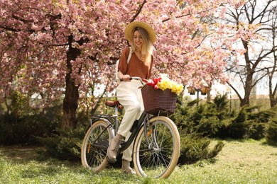 Beautiful young woman with bicycle and flowers in park on pleasant spring day