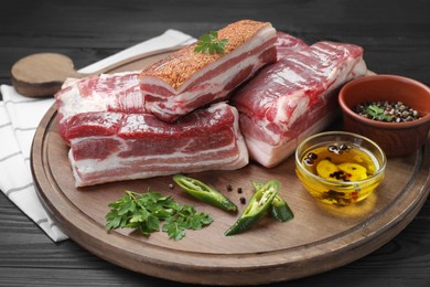 Photo of Pieces of raw pork belly, chili pepper, peppercorns, oil and parsley on black wooden table, closeup