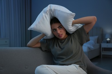 Photo of Unhappy young woman covering ears with pillow in living room at night. Noisy neighbours