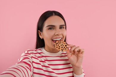 Young woman with chocolate chip cookie taking selfie on pink background