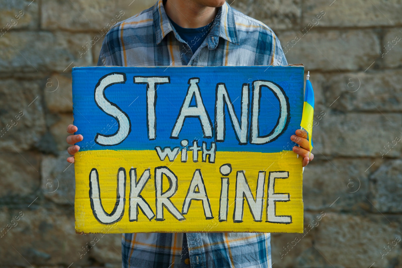 Photo of Woman holding poster in colors of national flag and words Stand With Ukraine against brick wall outdoors, closeup