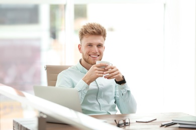 Portrait of handsome young man with cup of coffee at desk in office