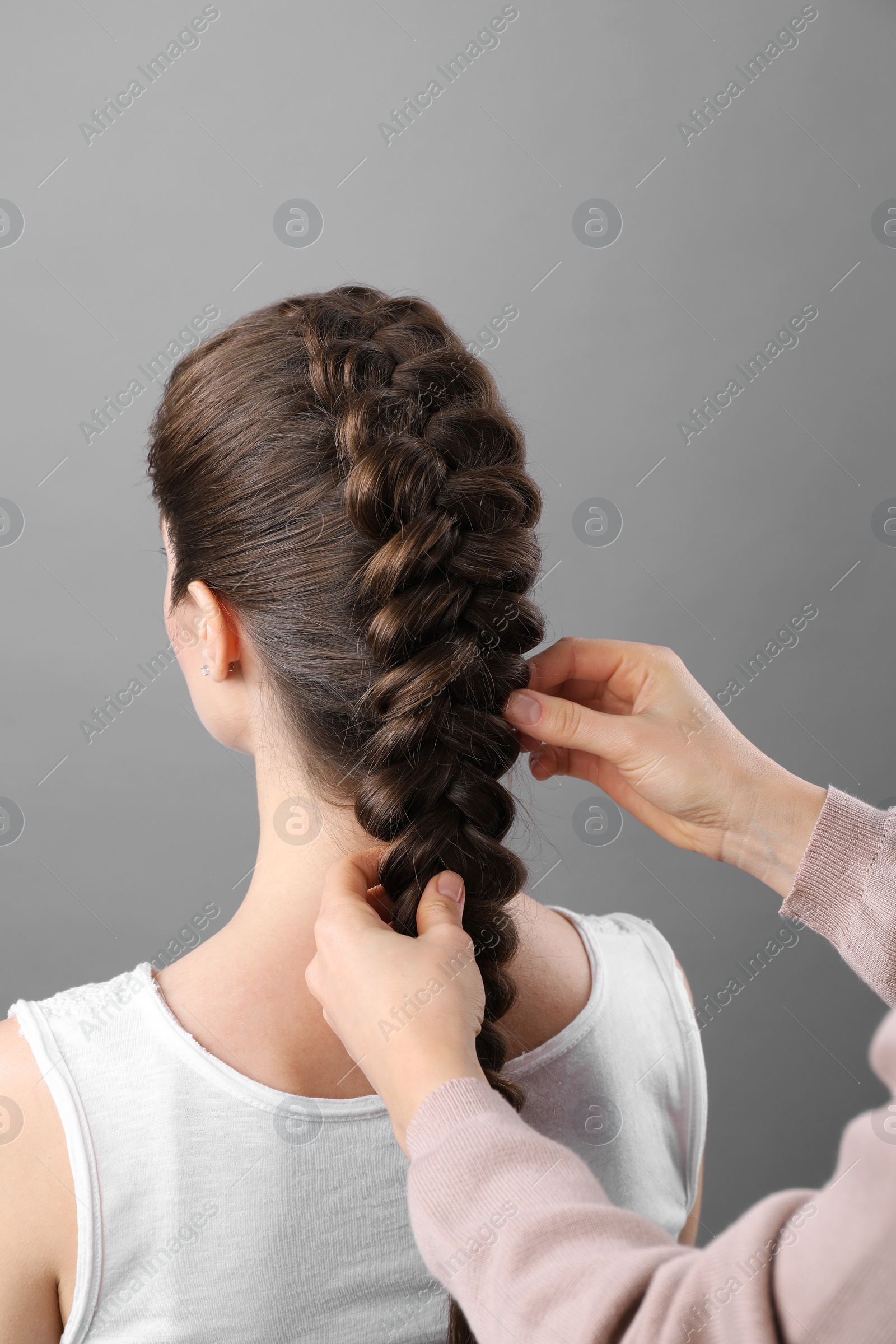 Photo of Professional stylist braiding woman's hair on grey background, closeup