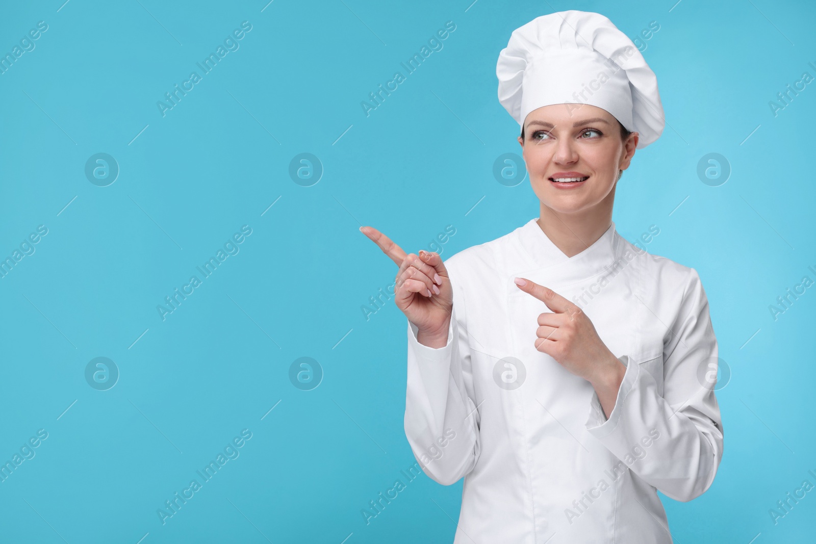 Photo of Happy woman chef in uniform pointing at something on light blue background, space for text