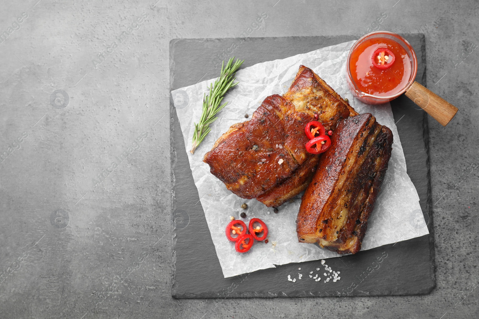 Photo of Pieces of baked pork belly served with sauce, rosemary and chili pepper on grey table, top view. Space for text