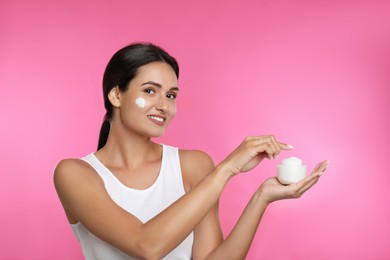 Young woman applying facial cream on pink background