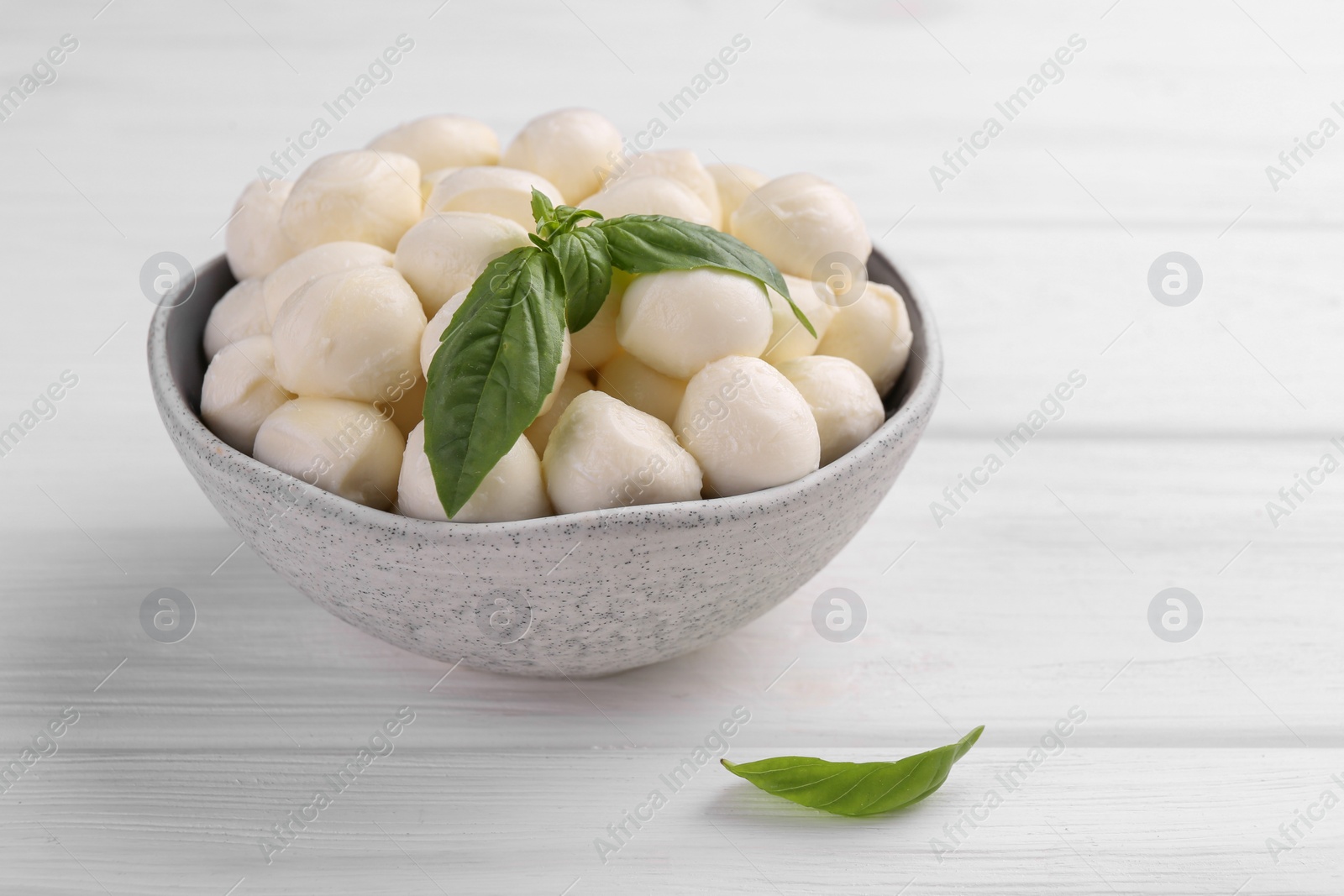 Photo of Tasty mozzarella balls and basil leaves in bowl on white wooden table, space for text