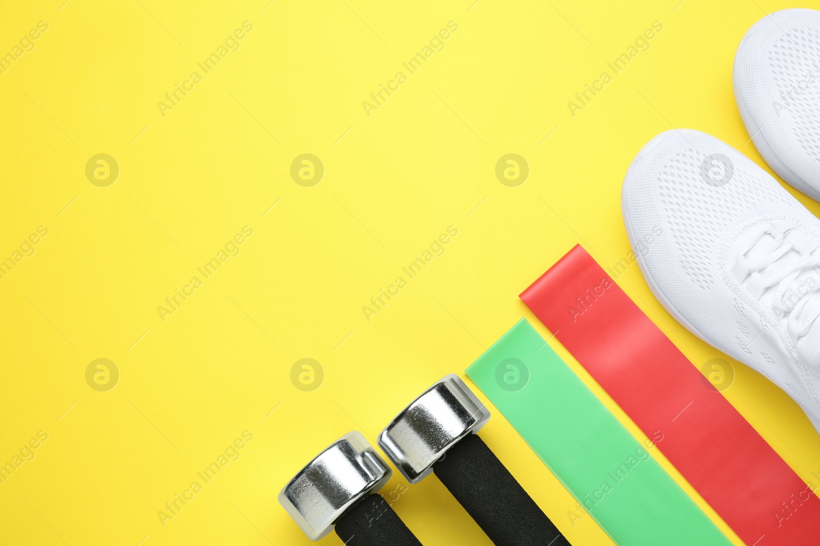 Photo of Different fitness elastic bands, sneakers and dumbbells on yellow background, flat lay. Space for text