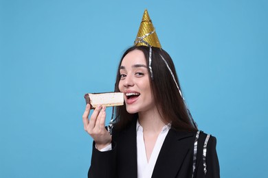 Photo of Woman in party hat eating piece of tasty cake on light blue background