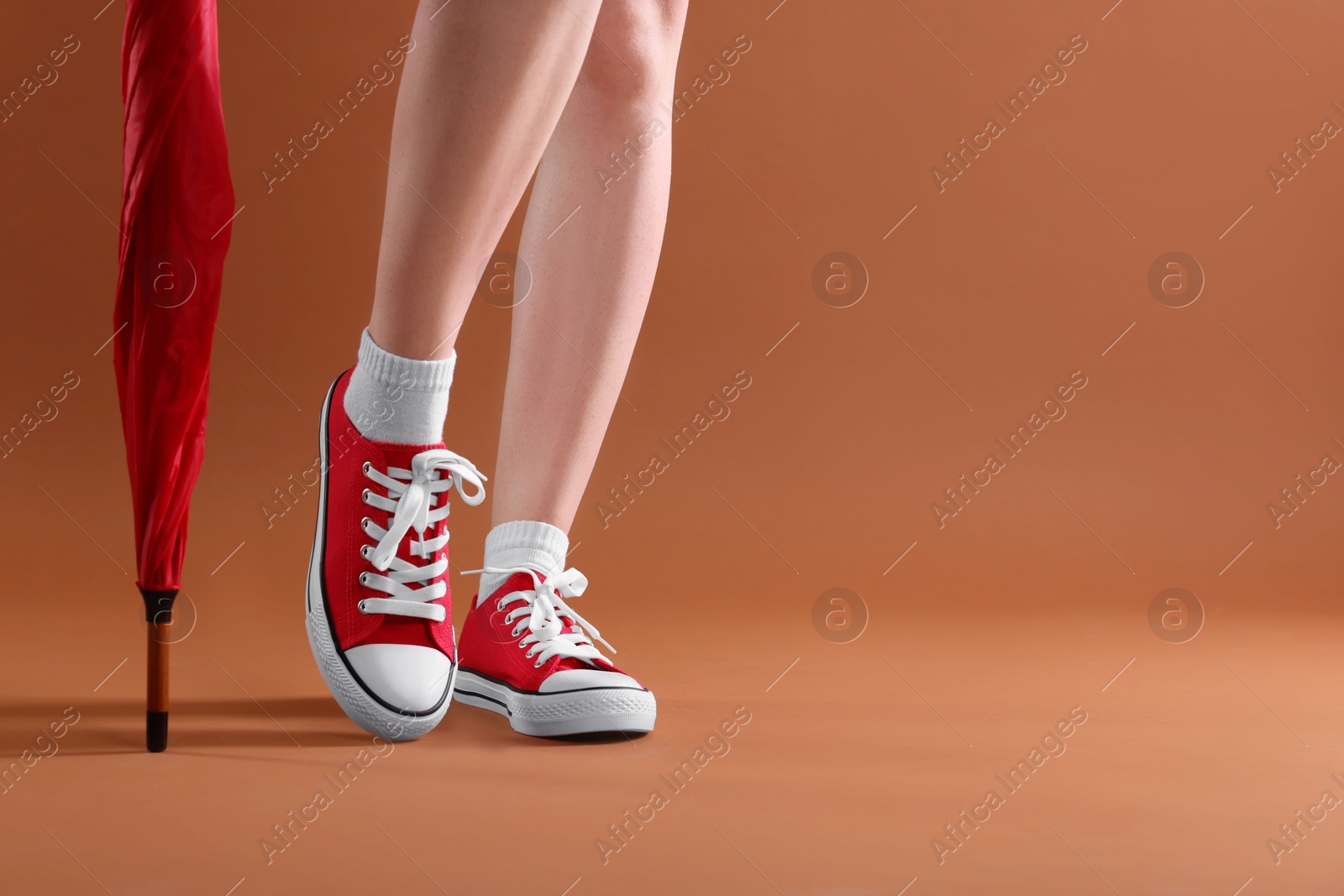 Photo of Woman in red classic old school sneakers and umbrella on brown background, closeup. Space for text