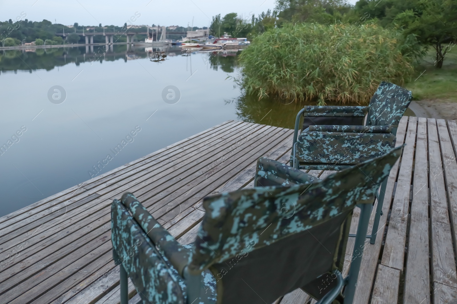 Photo of Camouflage fishing chairs on wooden pier near river, space for text