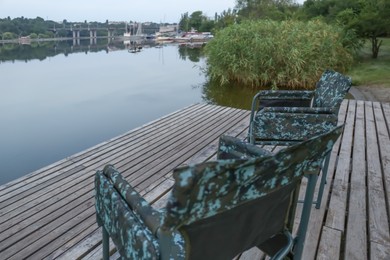 Camouflage fishing chairs on wooden pier near river, space for text