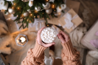 Woman with cup of delicious hot drink near Christmas tree at home, top view