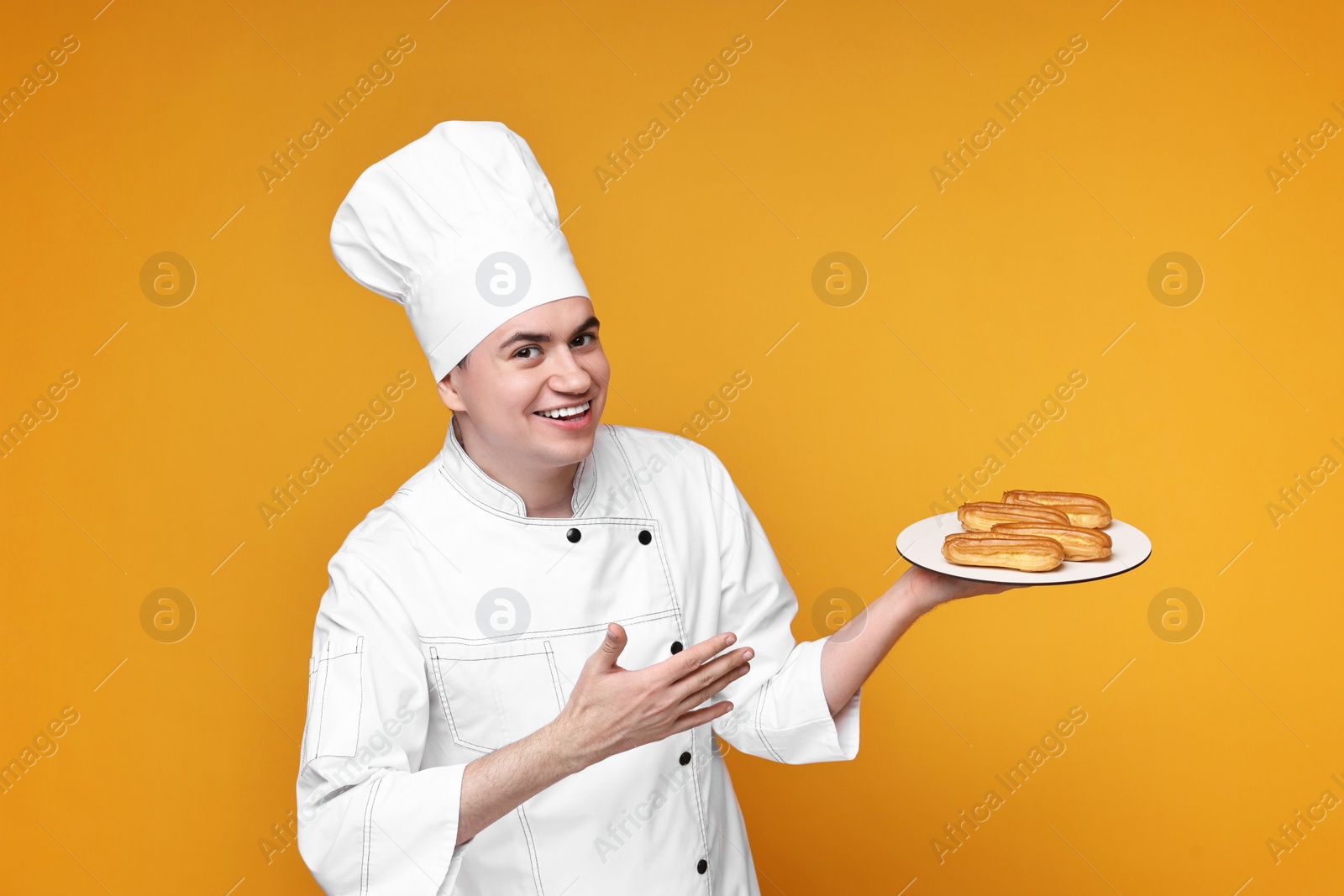 Photo of Portrait of happy confectioner in uniform holding plate with eclairs on orange background