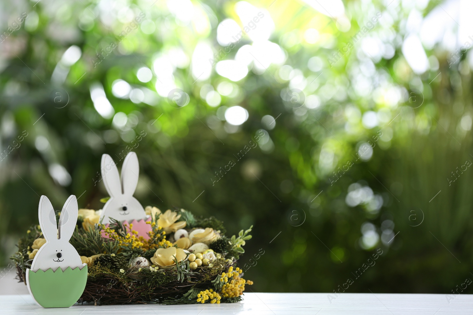 Photo of Decorative wreath and Easter bunny figures on white wooden table against blurred green background. Space for text